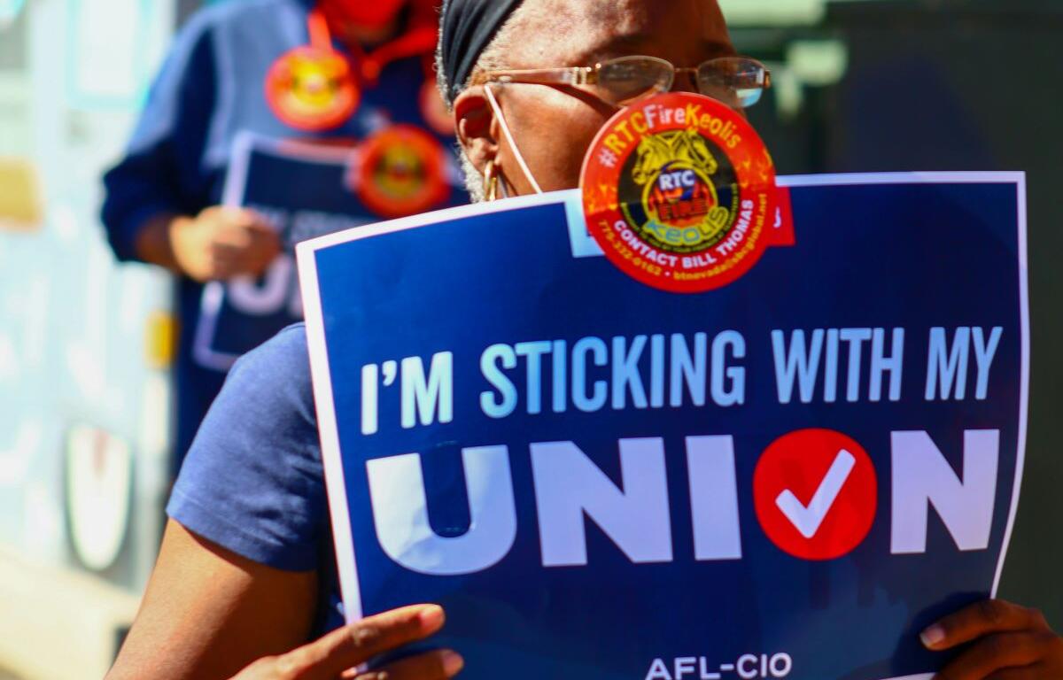 Protester holding a blue sign that reads "I'M STICKING WITH MY UNION" at a rally, wearing a mask and headband.