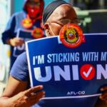 Protester holding a blue sign that reads "I'M STICKING WITH MY UNION" at a rally, wearing a mask and headband.