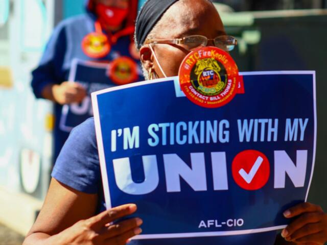 Protester holding a blue sign that reads "I'M STICKING WITH MY UNION" at a rally, wearing a mask and headband.