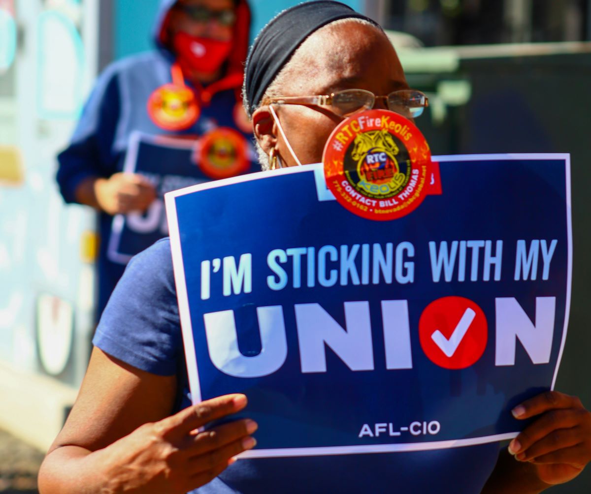Protester holding a blue sign that reads "I'M STICKING WITH MY UNION" at a rally, wearing a mask and headband.