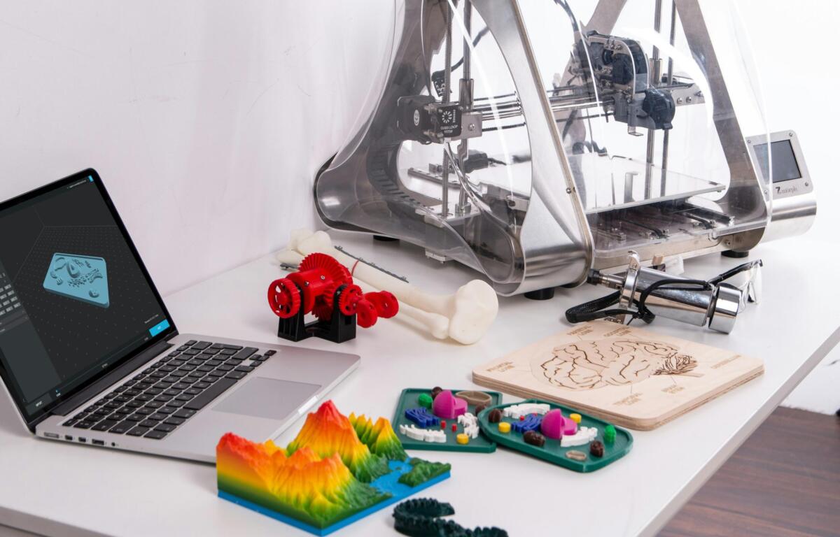 Laptop with a 3D dental model on screen, surrounded by colorful 3D-printed teeth and bone pieces and a silver 3D printer on a white desk