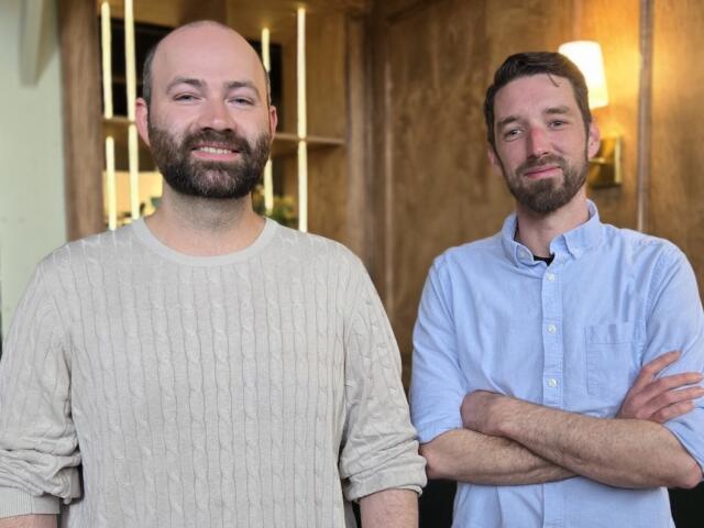 Two bearded men stand in a modern office, smiling at the camera.
