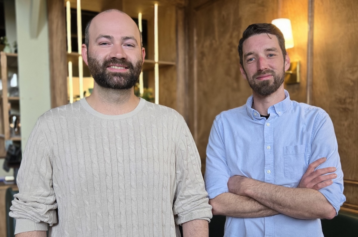 Two bearded men stand in a modern office, smiling at the camera.