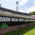 Stadium view with a large banner reading '2026 NCAA Men's Lacrosse Championships' along a brick wall and green field nearby, under a bright sky