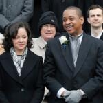 Smiling man in a dark suit with a white boutonniere, flanked by others in winter coats in a formal group photo.