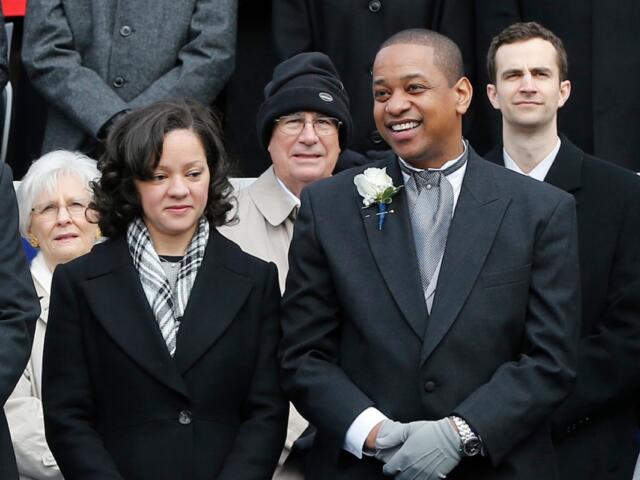 Smiling man in a dark suit with a white boutonniere, flanked by others in winter coats in a formal group photo.