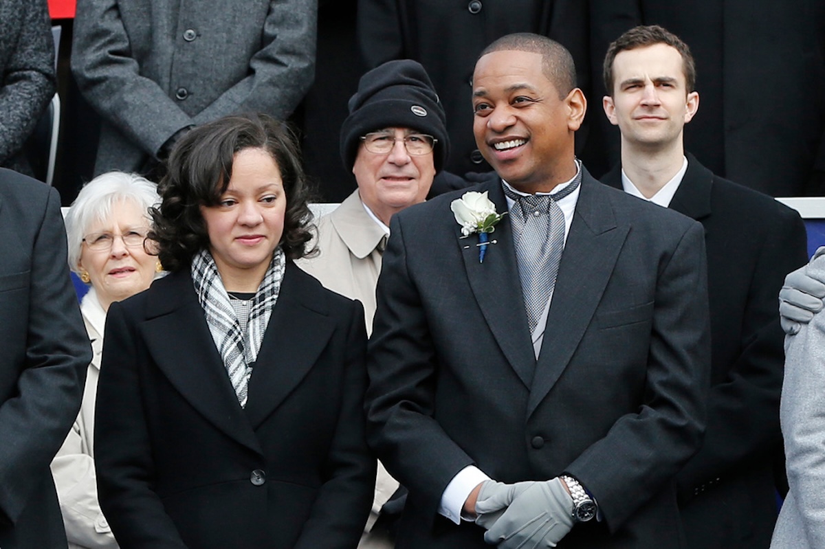 Smiling man in a dark suit with a white boutonniere, flanked by others in winter coats in a formal group photo.