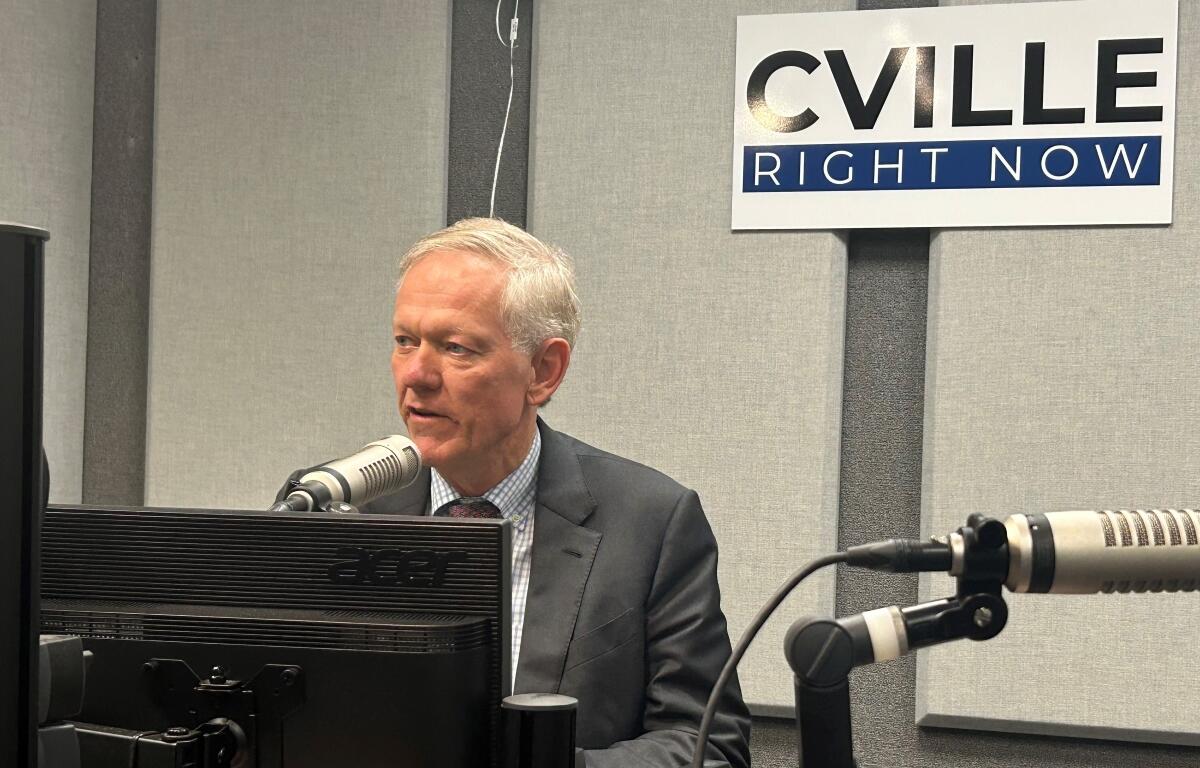 Man in a suit speaks into a studio microphone at a radio station, with a 'CVILLE RIGHT NOW' sign on the wall behind him.