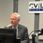 Man in a suit speaks into a studio microphone at a radio station, with a 'CVILLE RIGHT NOW' sign on the wall behind him.