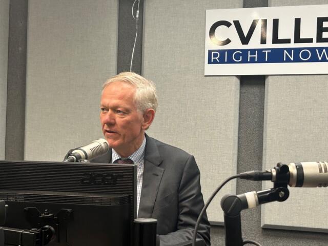 Man in a suit speaks into a studio microphone at a radio station, with a 'CVILLE RIGHT NOW' sign on the wall behind him.