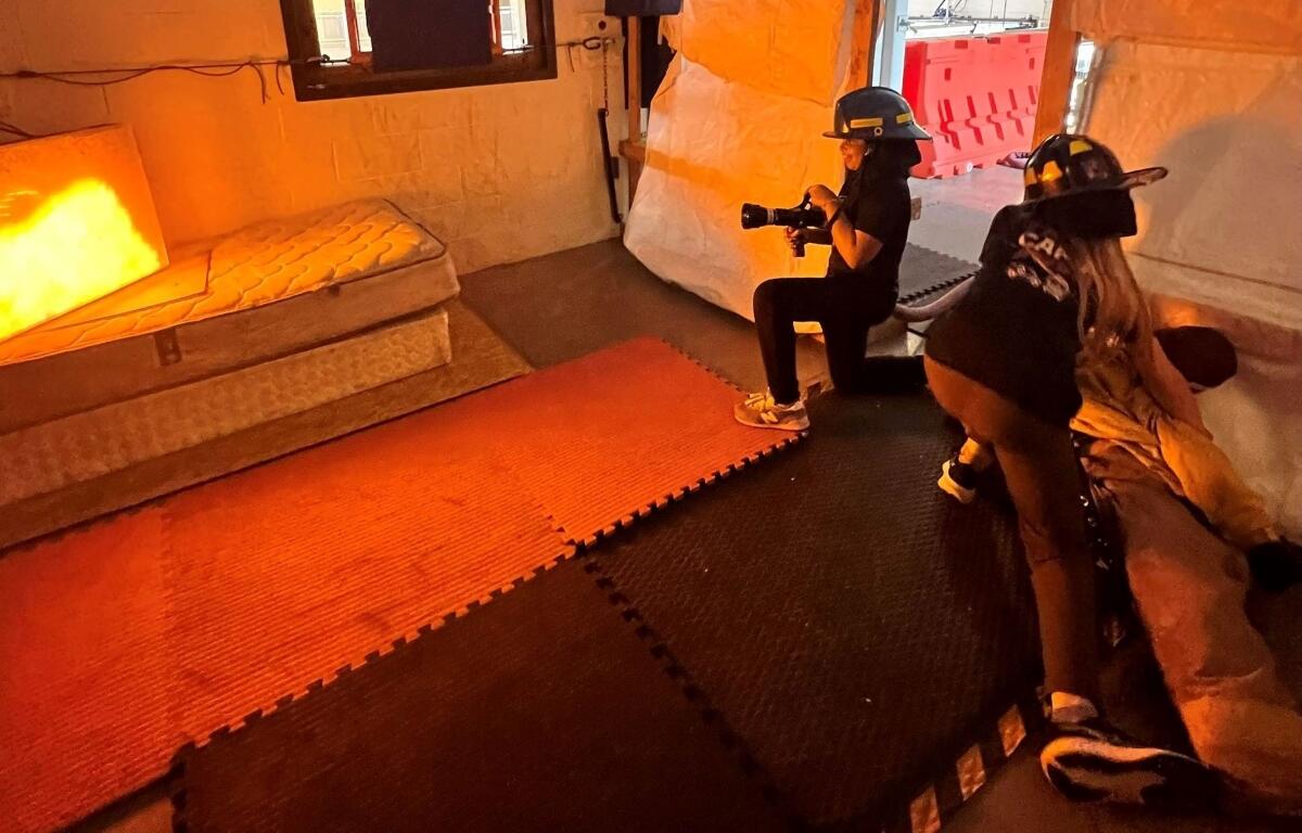 Two people wearing helmets film with a camera inside an orange-lit room on foam mats and a mattress nearby, resembling a set.