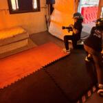 Two people wearing helmets film with a camera inside an orange-lit room on foam mats and a mattress nearby, resembling a set.