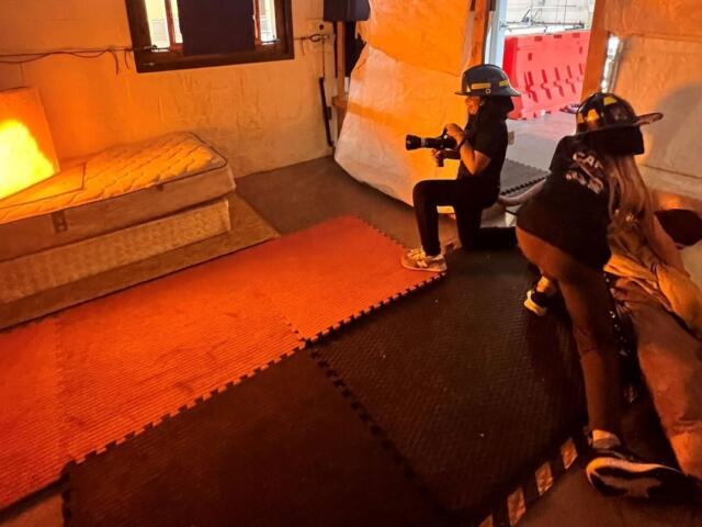 Two people wearing helmets film with a camera inside an orange-lit room on foam mats and a mattress nearby, resembling a set.