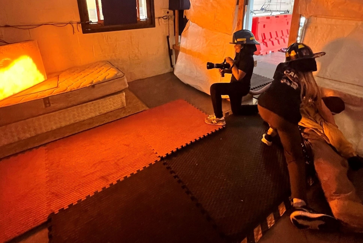 Two people wearing helmets film with a camera inside an orange-lit room on foam mats and a mattress nearby, resembling a set.