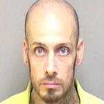 Mugshot of a bald man with a goatee, facing forward in a yellow shirt against a plain gray background, looking serious.”,