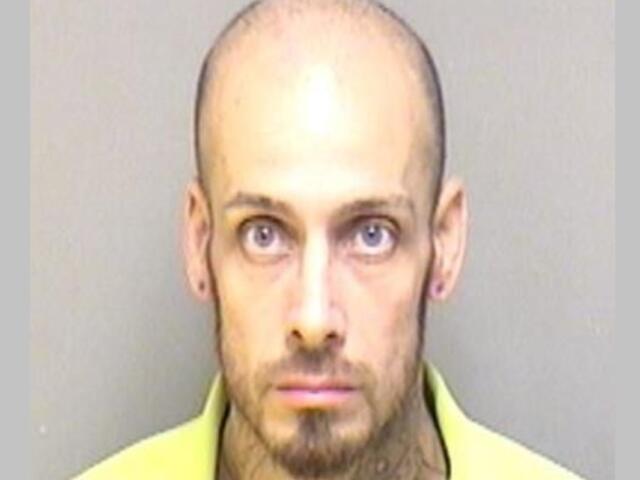 Mugshot of a bald man with a goatee, facing forward in a yellow shirt against a plain gray background, looking serious.”,