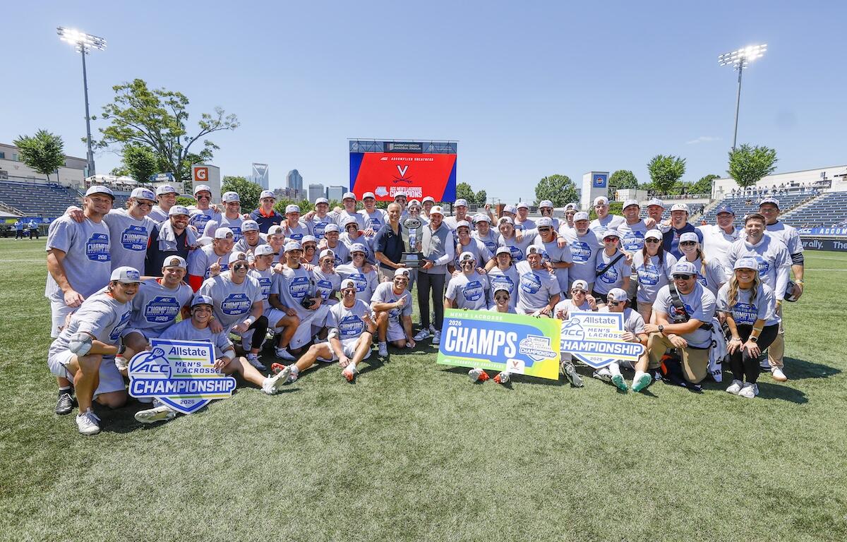 Large group of lacrosse players and staff pose on a sunny field around a tall trophy, with championship signs reading 'CHAMPS' and 'Allstate ACC Championship' in front of stadium seating.