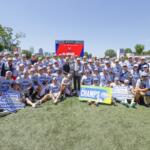 Large group of lacrosse players and staff pose on a sunny field around a tall trophy, with championship signs reading 'CHAMPS' and 'Allstate ACC Championship' in front of stadium seating.