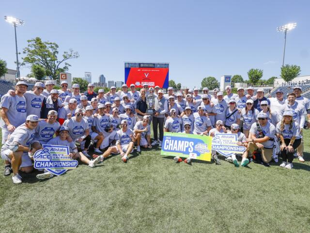 Large group of lacrosse players and staff pose on a sunny field around a tall trophy, with championship signs reading 'CHAMPS' and 'Allstate ACC Championship' in front of stadium seating.