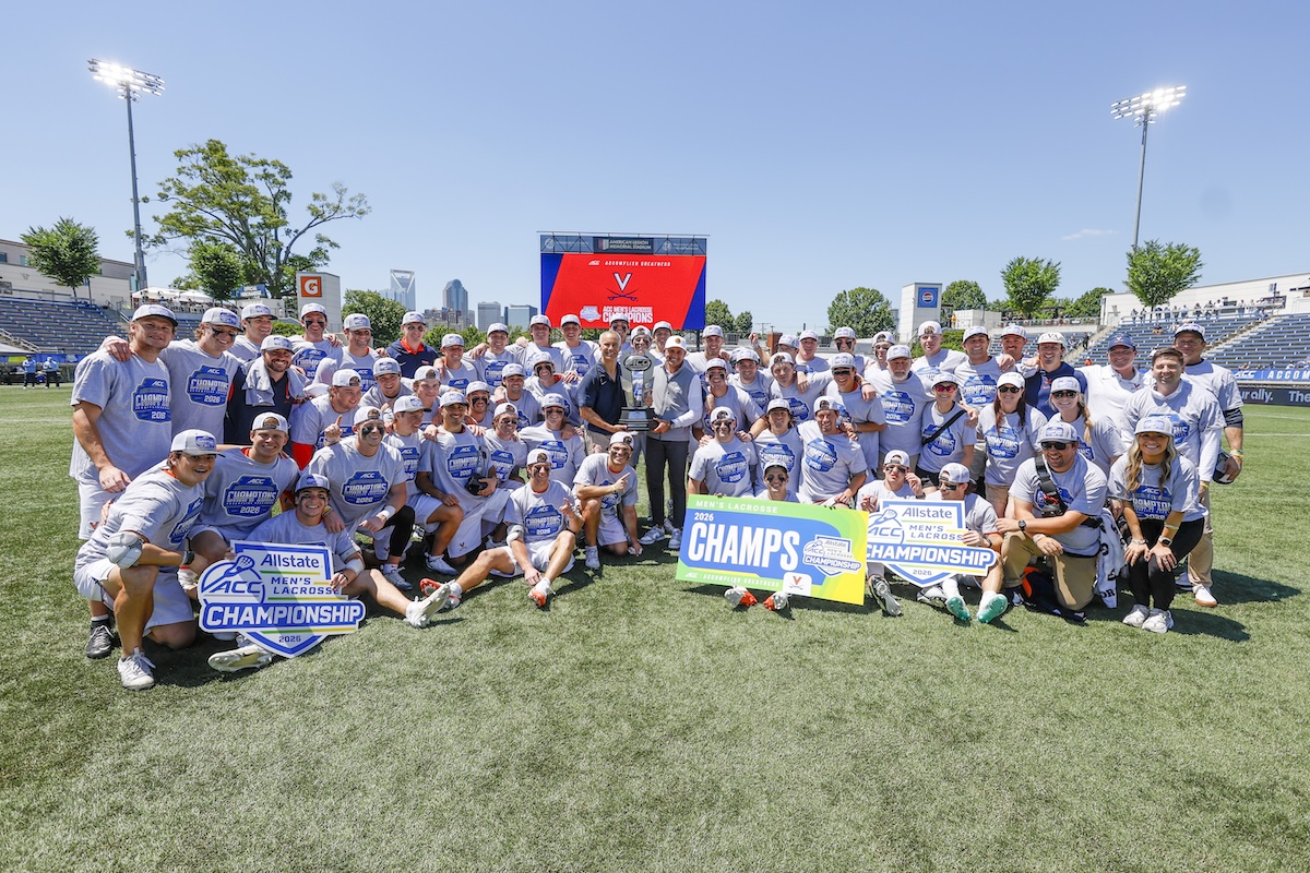 Large group of lacrosse players and staff pose on a sunny field around a tall trophy, with championship signs reading 'CHAMPS' and 'Allstate ACC Championship' in front of stadium seating.