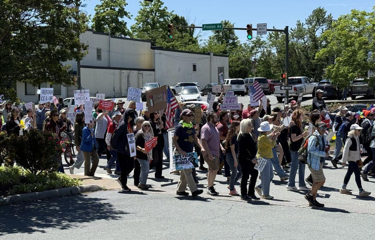 A group of diverse protesters marching across a street at a sunny intersection, holding handmade signs and some American flags.