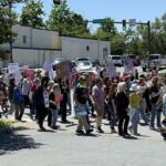 A group of diverse protesters marching across a street at a sunny intersection, holding handmade signs and some American flags.
