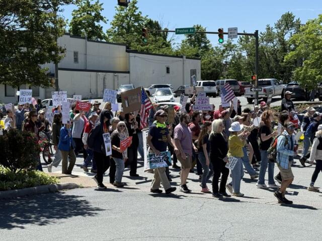 A group of diverse protesters marching across a street at a sunny intersection, holding handmade signs and some American flags.