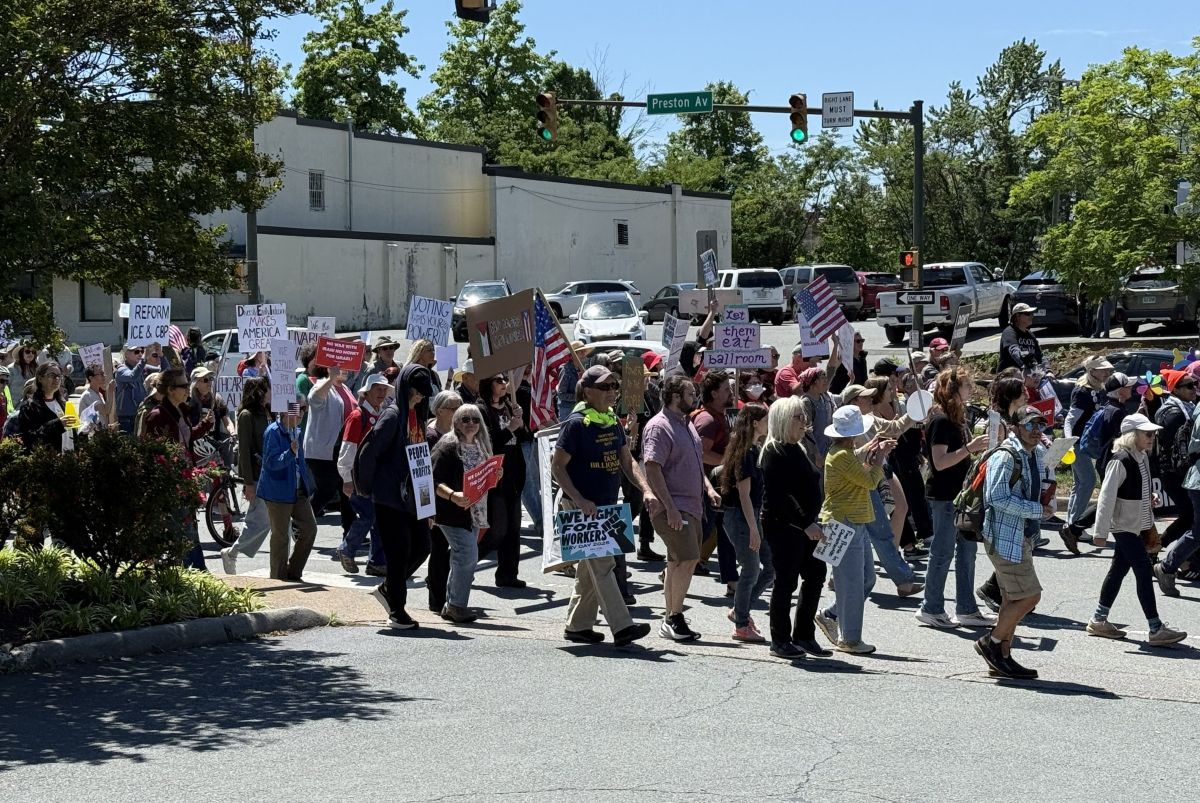 A group of diverse protesters marching across a street at a sunny intersection, holding handmade signs and some American flags.
