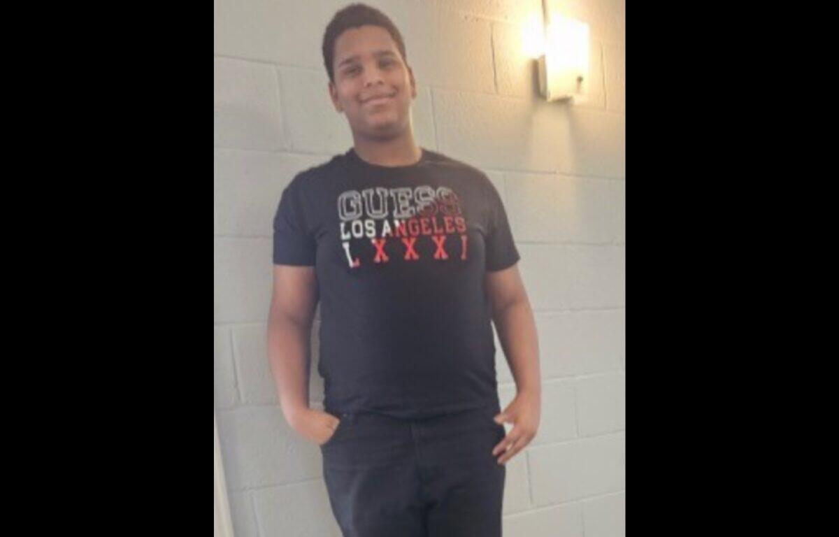 Young man standing with hands in pockets against a gray cinder block wall, wearing a black T-shirt.