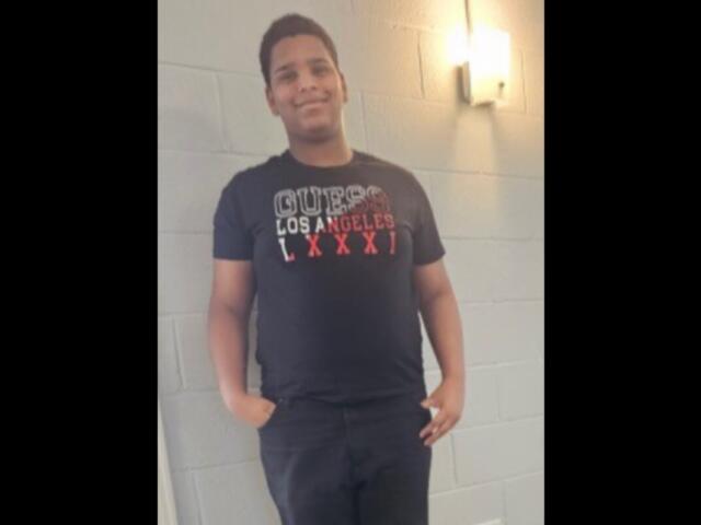 Young man standing with hands in pockets against a gray cinder block wall, wearing a black T-shirt.