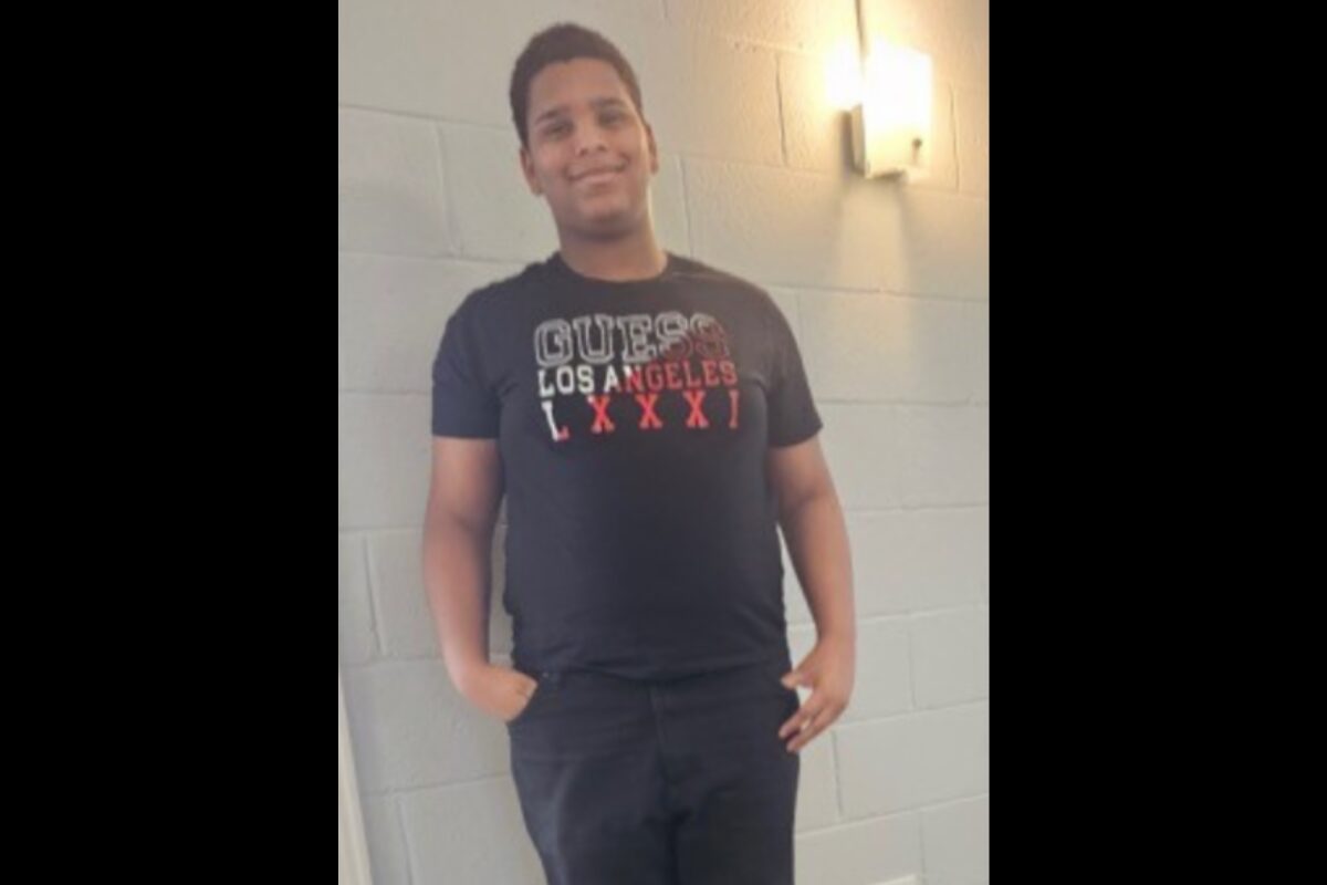 Young man standing with hands in pockets against a gray cinder block wall, wearing a black T-shirt.
