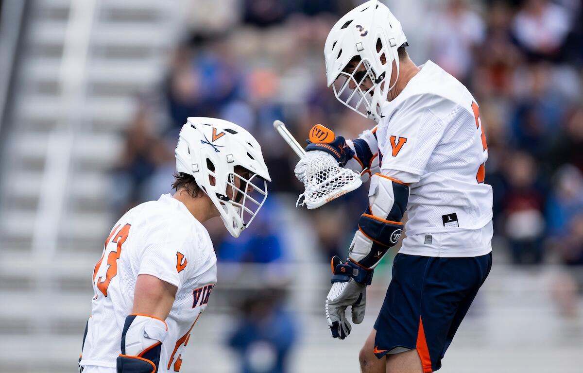 Two lacrosse players in white Virginia uniforms face off on a field, helmets on and sticks ready.