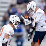 Two lacrosse players in white Virginia uniforms face off on a field, helmets on and sticks ready.
