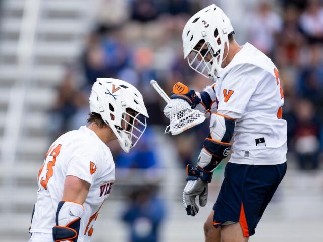 Two lacrosse players in white Virginia uniforms face off on a field, helmets on and sticks ready.