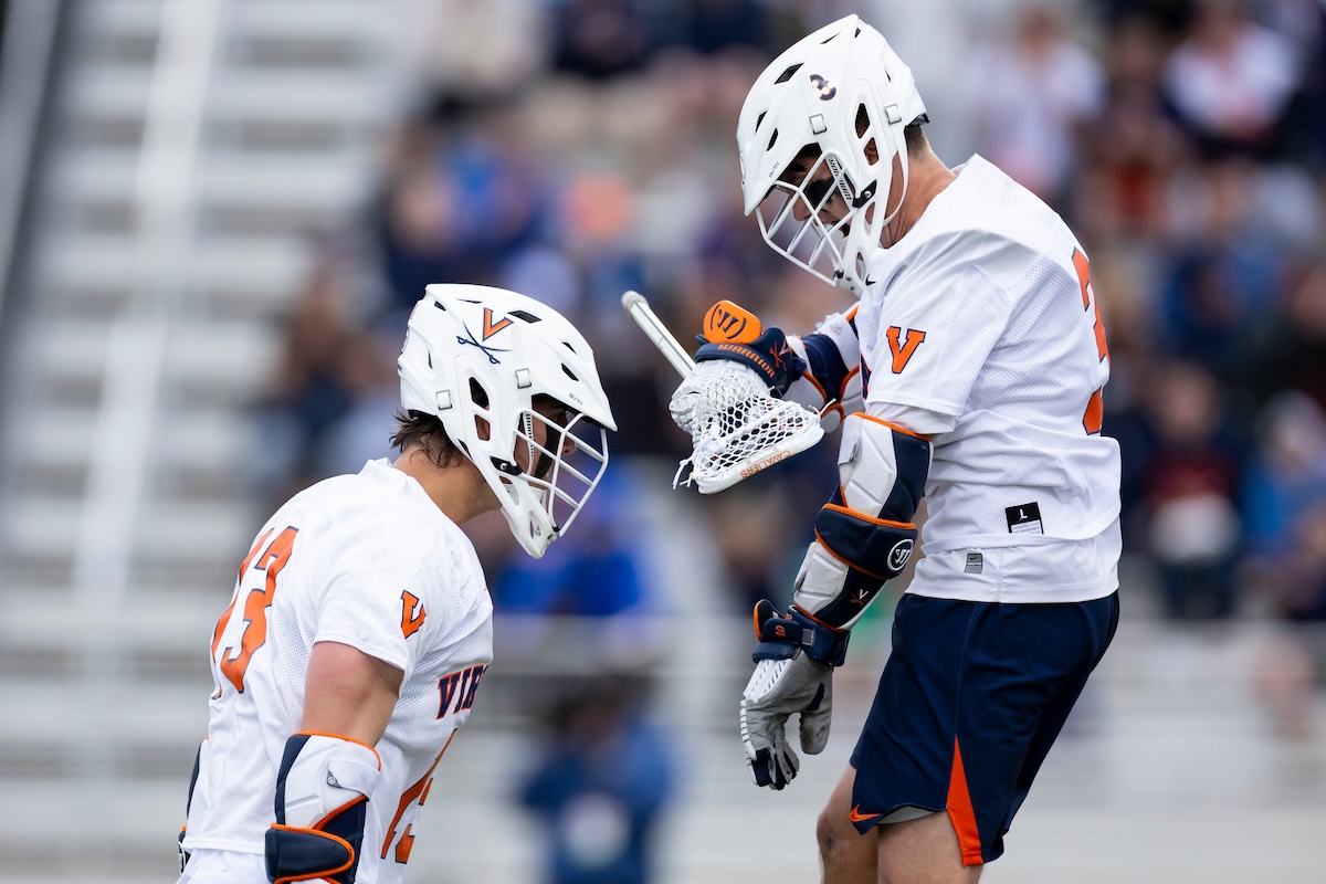Two lacrosse players in white Virginia uniforms face off on a field, helmets on and sticks ready.