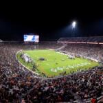 Nighttime football game in a packed outdoor stadium, field lit by tall floodlights with a large crowd in the stands.