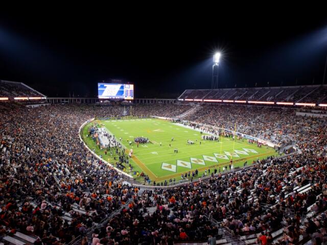 Nighttime football game in a packed outdoor stadium, field lit by tall floodlights with a large crowd in the stands.