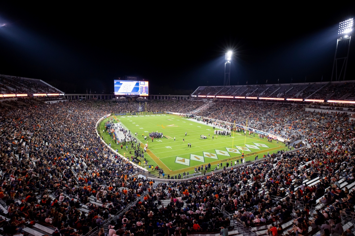 Nighttime football game in a packed outdoor stadium, field lit by tall floodlights with a large crowd in the stands.