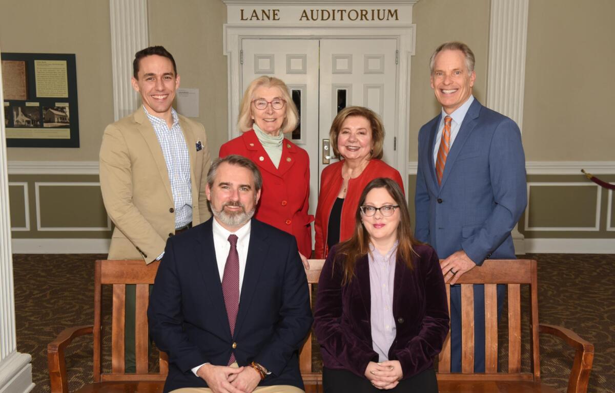 Six adults in business attire pose for a group photo in a hallway in front of a double-door entrance labeled 'Lane Auditorium' with a wood bench in the foreground.