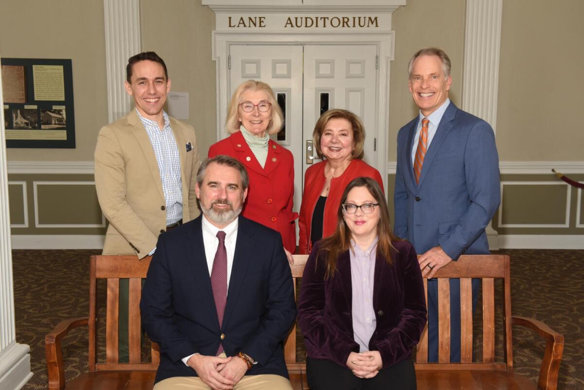 Six adults in business attire pose for a group photo in a hallway in front of a double-door entrance labeled 'Lane Auditorium' with a wood bench in the foreground.