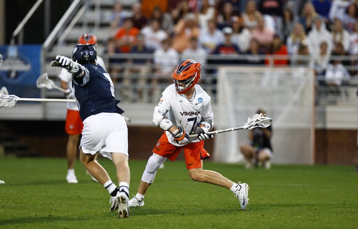 Virginia lacrosse player in white jersey and orange helmet dribbles the ball while dodging a defender on a green field with a crowded stands backdrop.