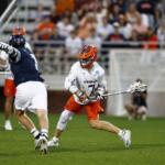 Virginia lacrosse player in white jersey and orange helmet dribbles the ball while dodging a defender on a green field with a crowded stands backdrop.