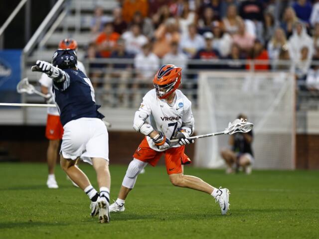 Virginia lacrosse player in white jersey and orange helmet dribbles the ball while dodging a defender on a green field with a crowded stands backdrop.