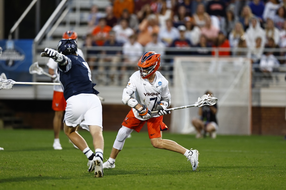 Virginia lacrosse player in white jersey and orange helmet dribbles the ball while dodging a defender on a green field with a crowded stands backdrop.