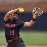 Softball pitcher in black uniform labeled HOOS, number 13, throws a yellow ball with glove raised mid-throw on a field.