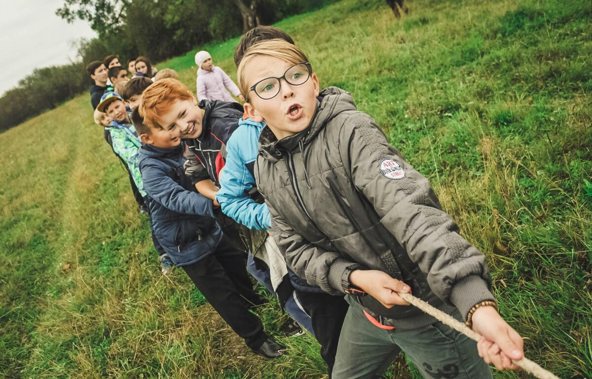 Group of children outdoors on a grassy field, lined up to tug a rope; foreground boy wearing glasses and a gray jacket pulls the rope toward the camera.