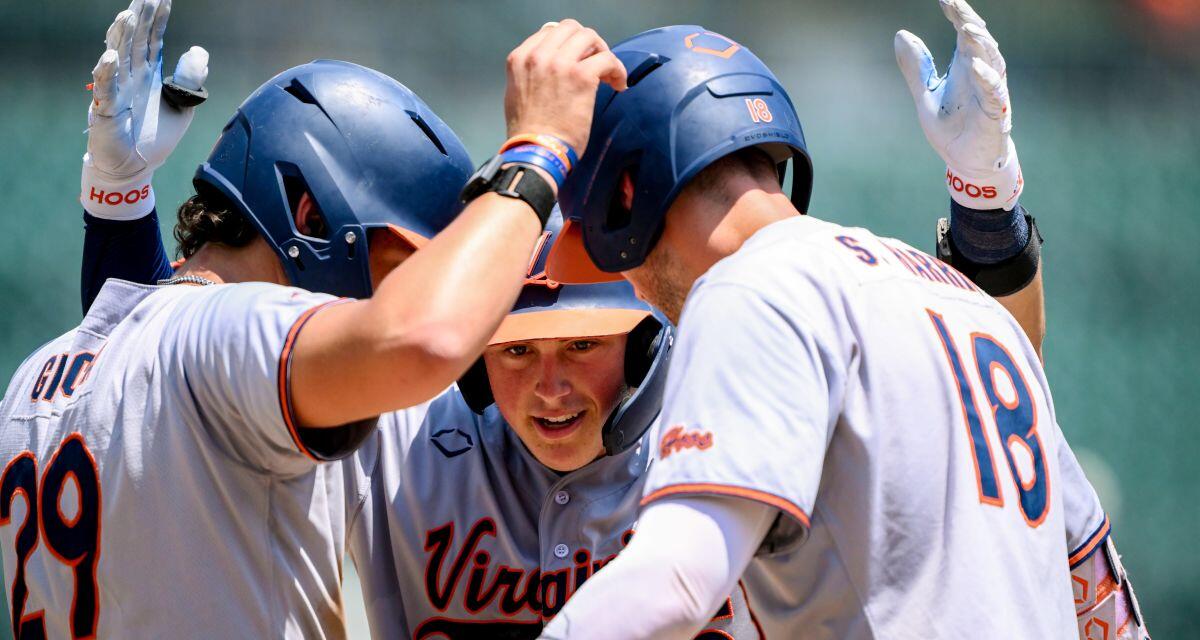 Virginia Cavaliers baseball players in white uniforms celebrate in a huddle on the field, high-fiving each other around a central player.