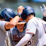 Virginia Cavaliers baseball players in white uniforms celebrate in a huddle on the field, high-fiving each other around a central player.