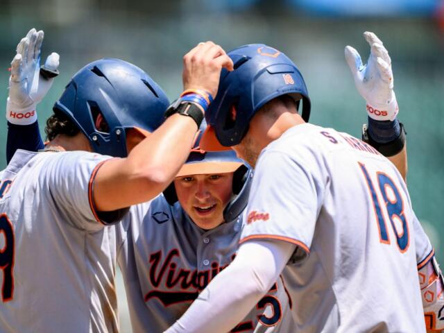 Virginia Cavaliers baseball players in white uniforms celebrate in a huddle on the field, high-fiving each other around a central player.