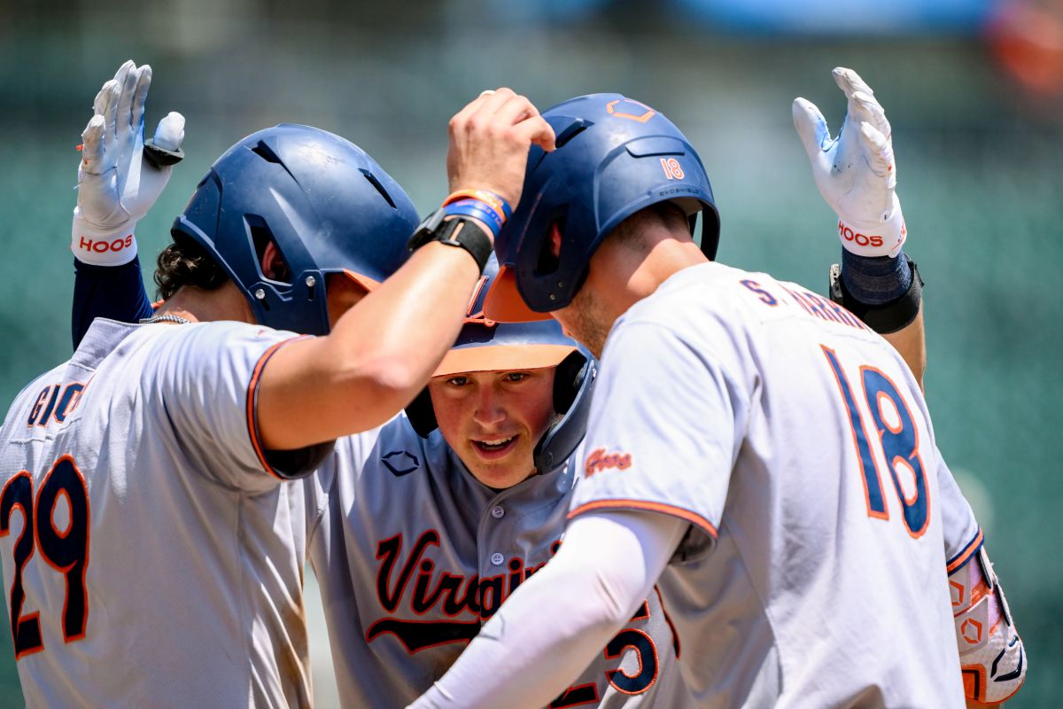 Virginia Cavaliers baseball players in white uniforms celebrate in a huddle on the field, high-fiving each other around a central player.